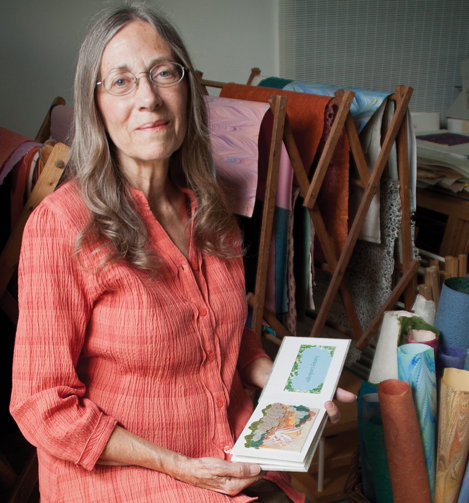 Diane Maurer holding a small booklet of her artwork and standing in front of some rolls of paper.